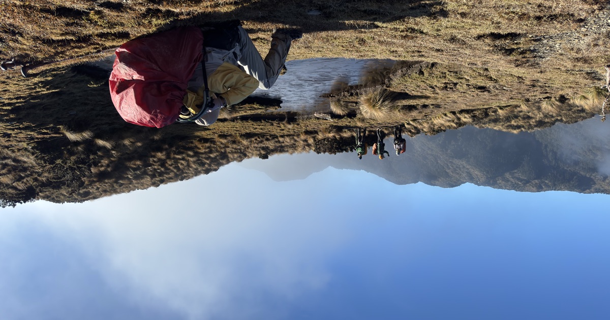登山者背著背包在高山池畔休息，遠方是雲霧繚繞的山巒